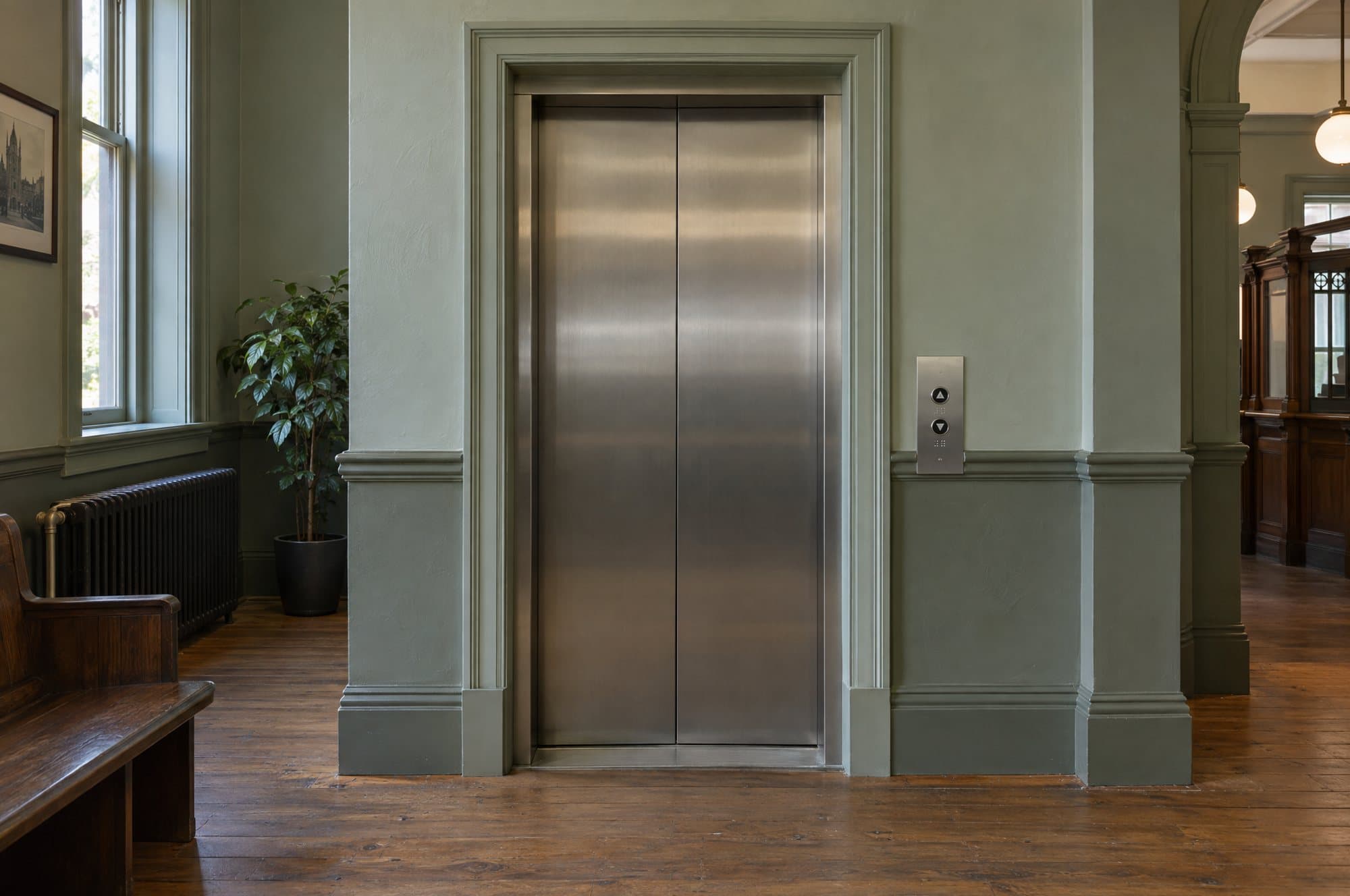 The lift in the counter hall: brushed steel doors set in a grey-green wall, with up and down buttons and braille at the side.