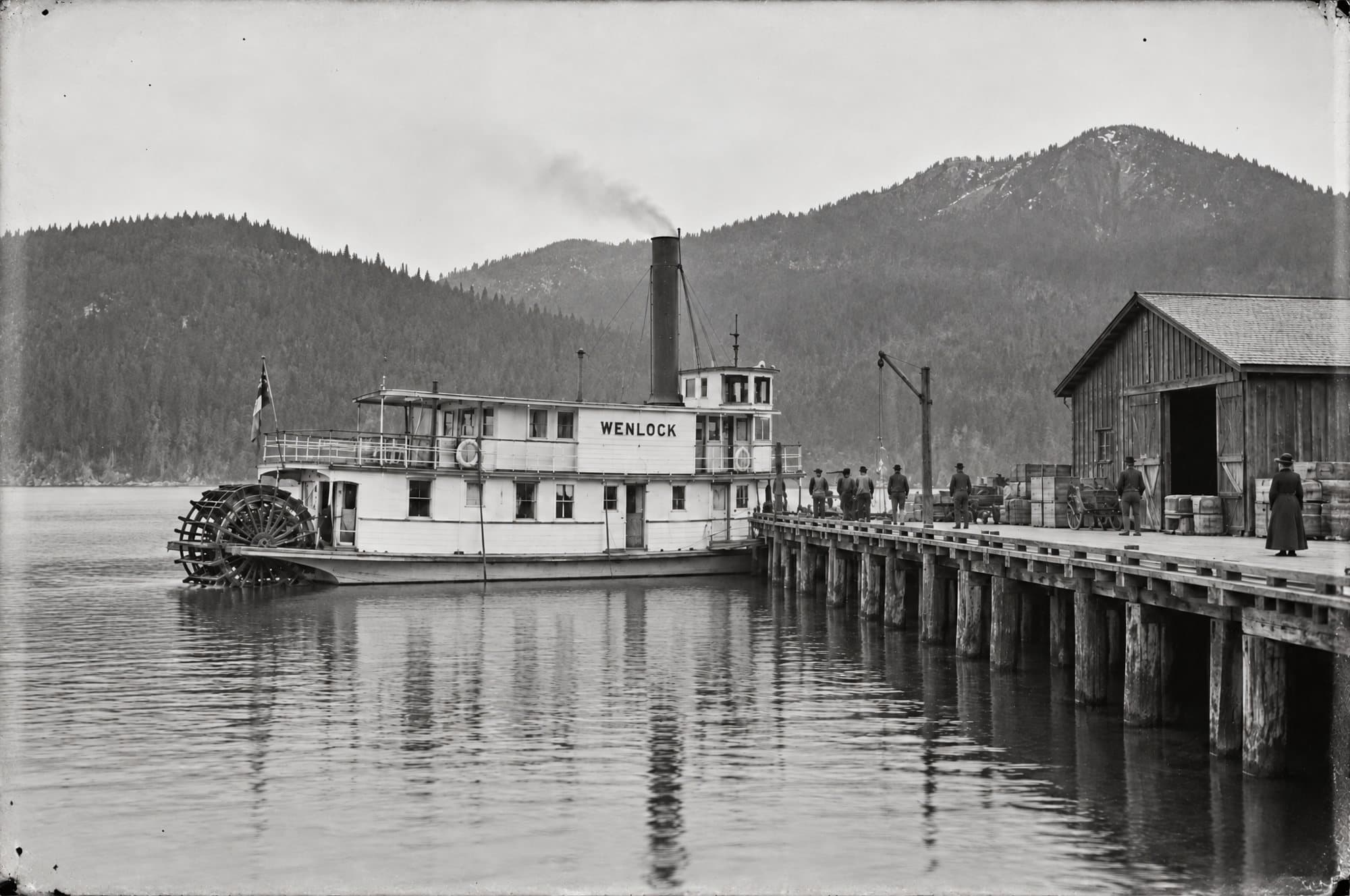 The sternwheeler SS Wenlock tied up at the town wharf, smoke from her stack, men and crates on the dock beside a wooden freight shed, forested hills behind.