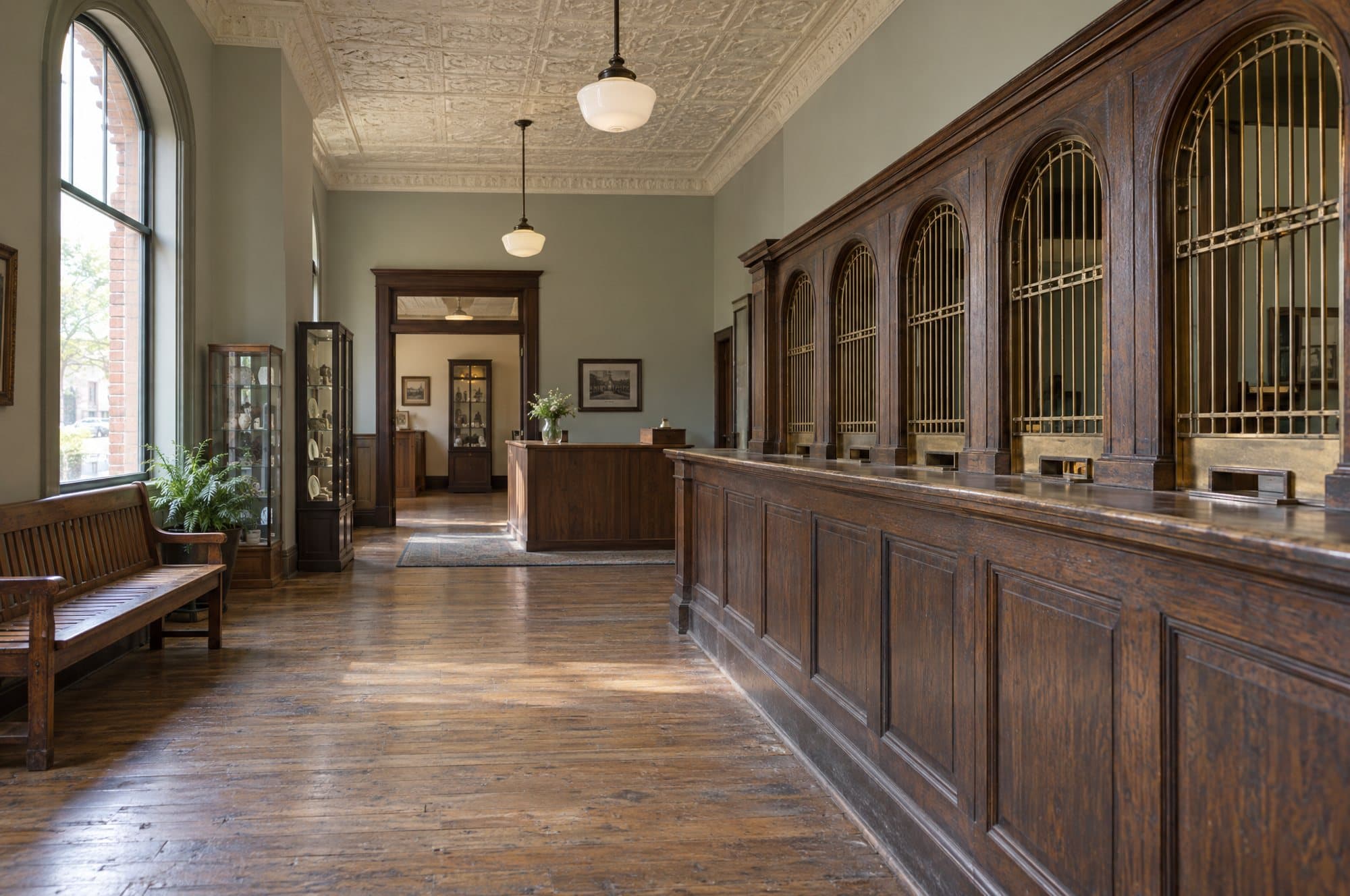 The counter hall inside the museum: a long oak counter with brass-barred wicket windows, worn fir floorboards, a wooden bench and tall arched windows.