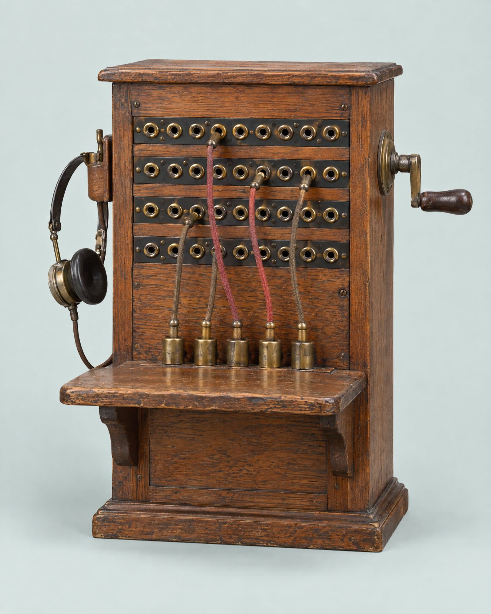 An oak telephone switchboard with rows of brass jacks, red patch cords, a crank and a headset.