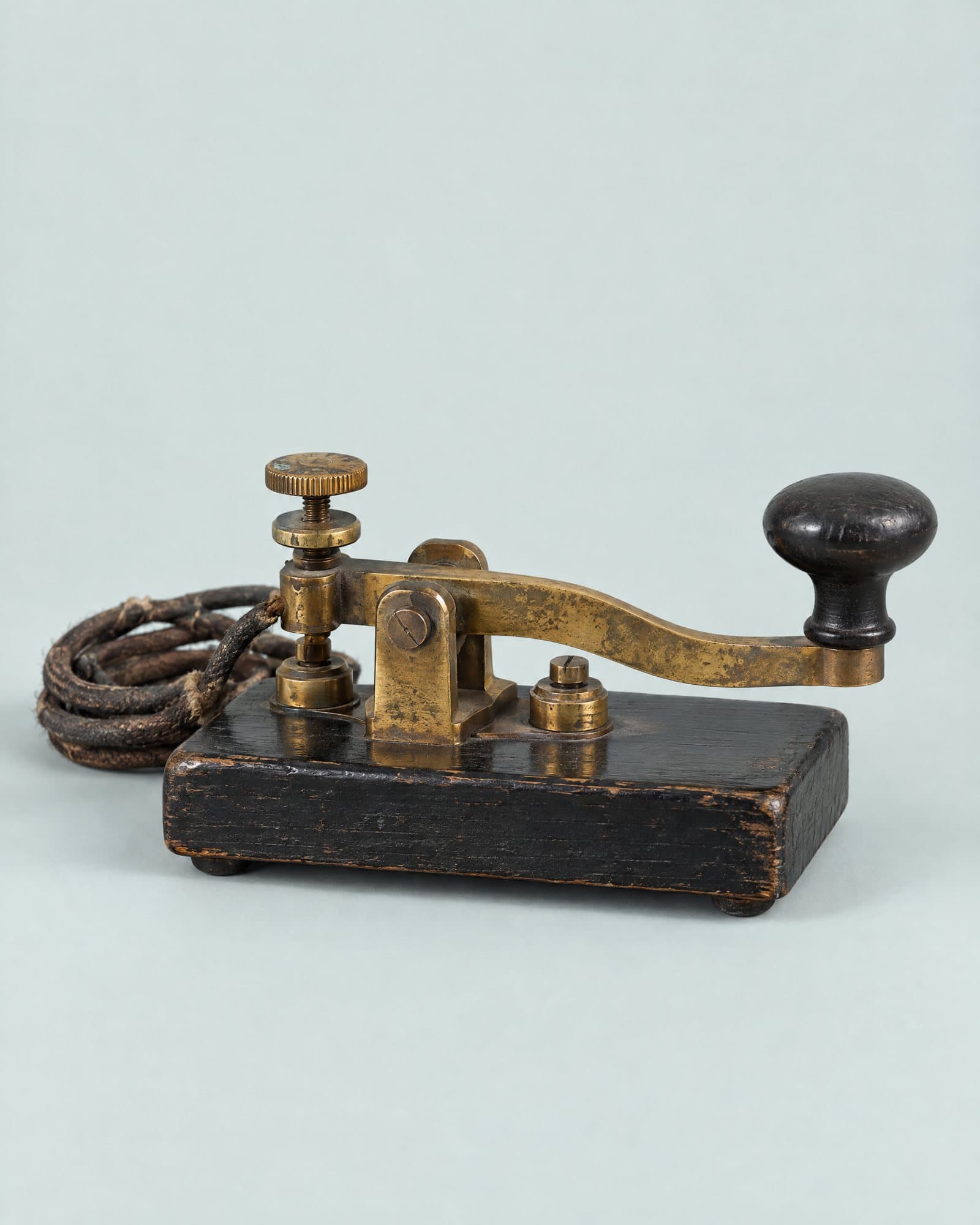 A brass telegraph key with a black knob, mounted on a black wooden base with a braided cord.